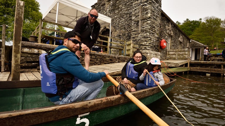 A family sat in a traditional wooden rowboat next to a stone jetty with a stone boathouse in the background.